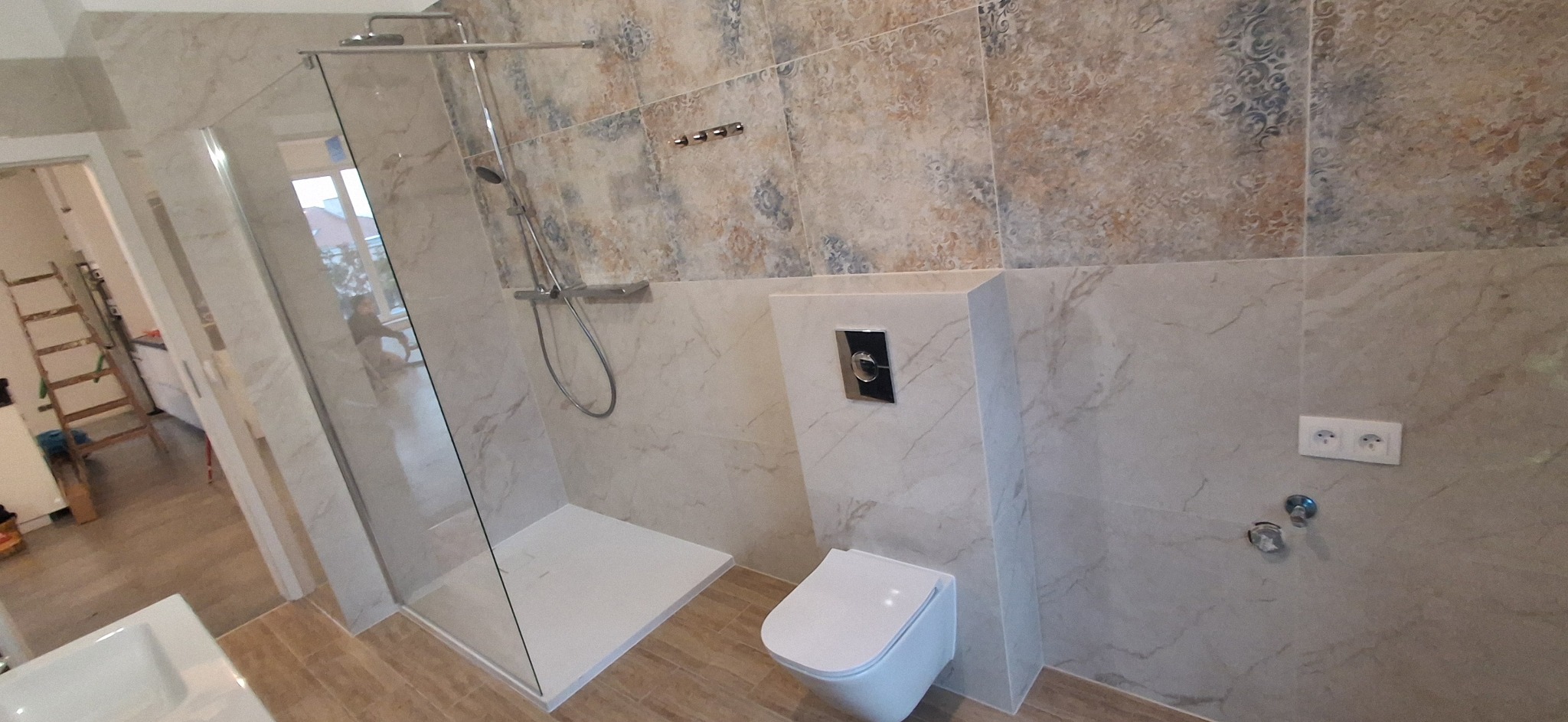 Wykończona łazienka z kabiną prysznicową typu walk-in, wiszącą toaletą i płytkami w odcieniach beżu i brązu. Widoczna drabina w tle sugeruje trwające prace.