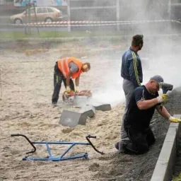 Pracownicy budowlani podczas układania obrzeży chodnika z kostki granitowej, jeden tnie kostkę szlifierką kątową, drugi przecina kostki piłą spalinową w pomarańczowej kamizelce odblaskowej, w tle...