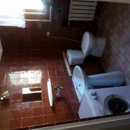 Ustawiona pod kątem fotografia łazienki z brązowymi płytkami, ukazująca toaletę, umywalkę na postumencie, pralkę i kaloryfer pod oknem.