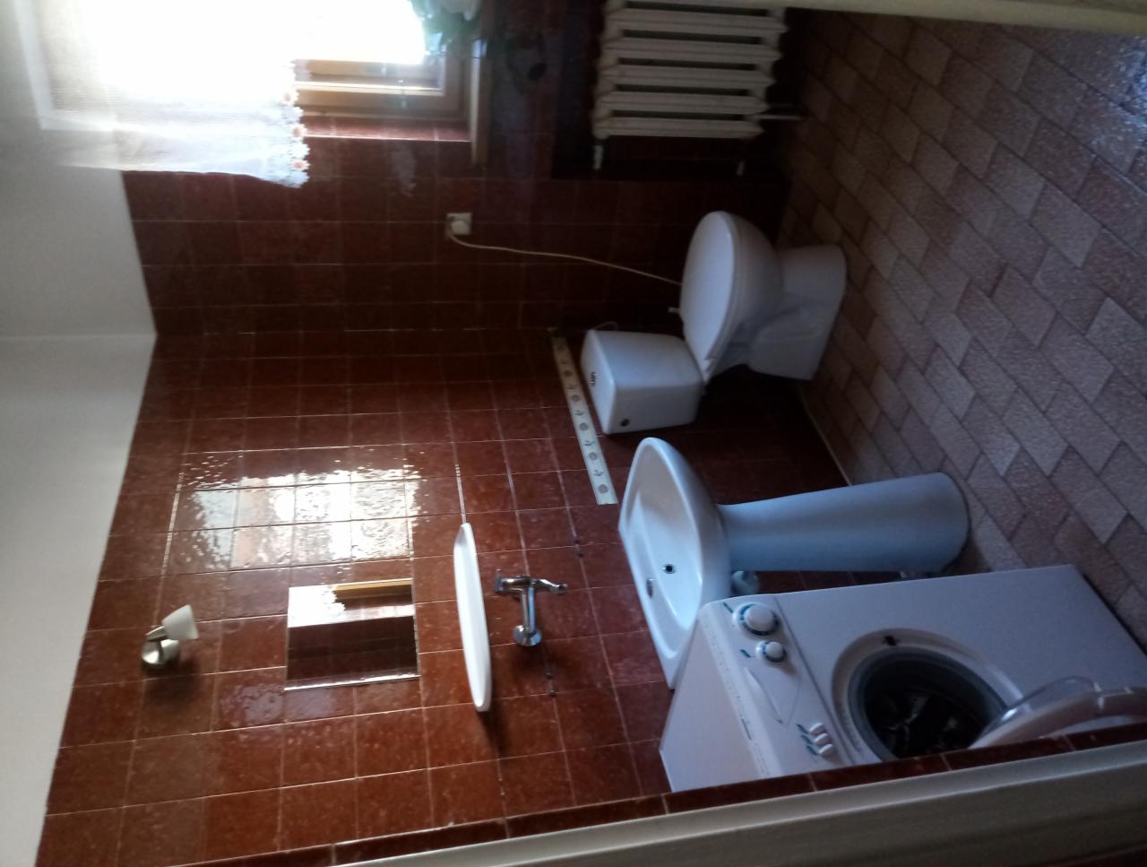 Ustawiona pod kątem fotografia łazienki z brązowymi płytkami, ukazująca toaletę, umywalkę na postumencie, pralkę i kaloryfer pod oknem.