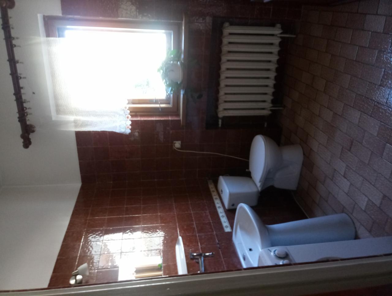 Wnętrze łazienki z brązowymi płytkami na ścianach i podłodze, biała umywalka i toaleta, grzejnik pod oknem z firanką i rośliną doniczkową.