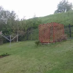 Drewniana ażurowa konstrukcja altany w ogrodzie na tle zielonego wzgórza, obok suszarka na pranie i grządki pod folią.