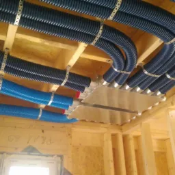 Niebieskie i granatowe rury wentylacyjne, mocowane metalowymi obejmami do drewnianej konstrukcji stropu, z widocznym metalowym rozdzielaczem powietrza.