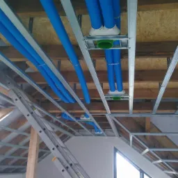 Montaż instalacji rekuperacji pod stropem poddasza z widocznymi niebieskimi rurami wentylacyjnymi, elementami mocującymi i drabina oparta o ścianę.