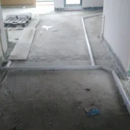 Surowe wnętrze w trakcie instalacji rur systemu ogrzewania podłogowego, widoczne białe rury ułożone na szarej, betonowej posadzce, ściany w stanie surowym, wiadro z zaprawą w rogu.