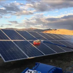 Czyszczenie paneli słonecznych za pomocą szczotki na wysięgniku, w tle wzg&oacute;rza i zachmurzone niebo.