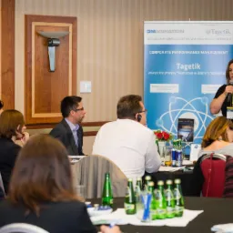 Kobieta z mikrofonem prowadzi prezentację dla grupy osób siedzących przy stole, za nią widoczne banery reklamowe z logo firm i hasłami dotyczącymi zarządzania finansami.