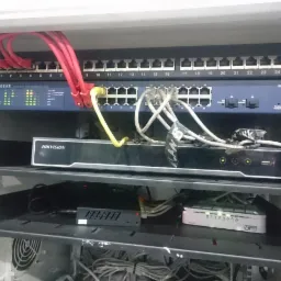 Szafa rack z zainstalowanym switchem Netgear GS724T, rejestratorem Hikvision i okablowaniem sieciowym, widoczne porty ethernetowe i diody sygnalizacyjne, na dolnej półce splątane kable.