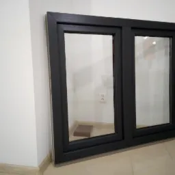 Dwukomorowe okno z czarną ramą, oparte o ścianę, z widocznym fragmentem podłogi i gniazdkiem elektrycznym w tle.