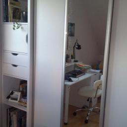 Xxx - Biała szafa przesuwna z lustrzanymi drzwiami, widoczny regał z książkami i biurko z lampką w odbiciu lustra.