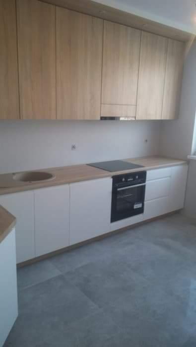 Jasna kuchnia z drewnianymi szafkami górnymi i białymi szafkami dolnymi, z wbudowaną płytą grzewczą i piekarnikiem, widok na szare płytki podłogowe.