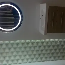 Nowoczesna łazienka z okrągłym lustrem z podświetleniem LED, umywalką z szafką wiszącą i geometryczną mozaiką na ścianie, uzupełniona płytkami podłogowymi w odcieniach beżu i brązu.