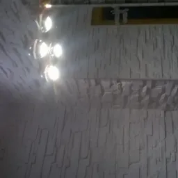 Ściana z dekoracyjną, białą okładziną imitującą kamień, oświetlona przez reflektory sufitowe, widoczny fragment okna z ramą w kolorze drewna.