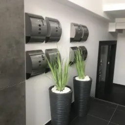 Szare skrzynki pocztowe z numerami 1, 2 i 5 na ścianie w korytarzu, obok donice z wysokimi trawami ozdobnymi i białymi kamieniami, ciemna podłoga i drzwi.