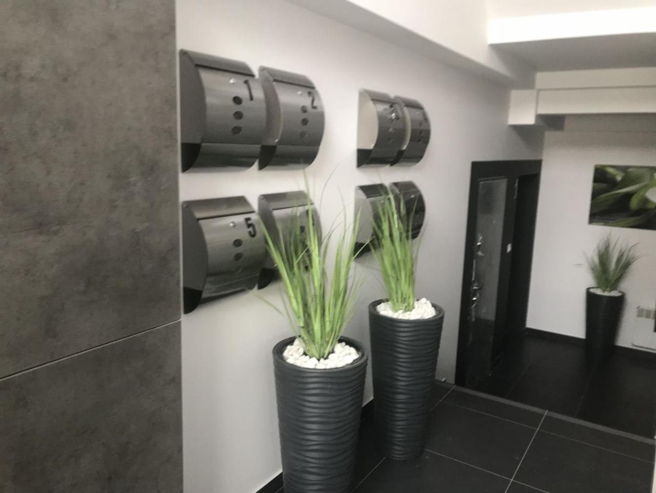 Szare skrzynki pocztowe z numerami 1, 2 i 5 na ścianie w korytarzu, obok donice z wysokimi trawami ozdobnymi i białymi kamieniami, ciemna podłoga i drzwi.