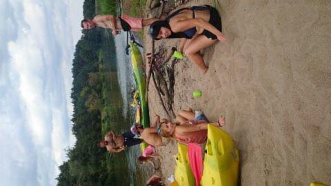 Grupa osób odpoczywa na piaszczystej plaży obok rzeki i kajaków, część osób w strojach kąpielowych, widoczne drzewa w tle.