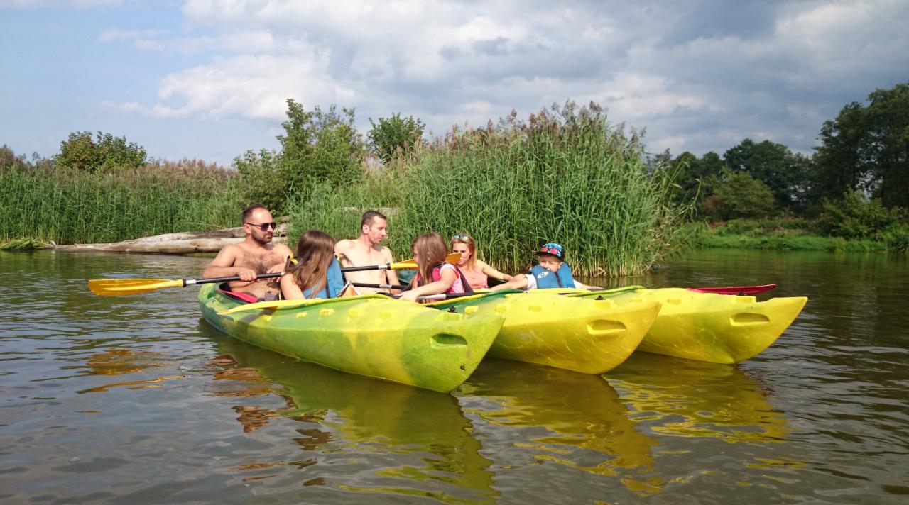 Grupa osób płynie trzema żółtymi kajakami po spokojnej rzece, otoczonej zieloną roślinnością i drzewami pod pochmurnym niebem.