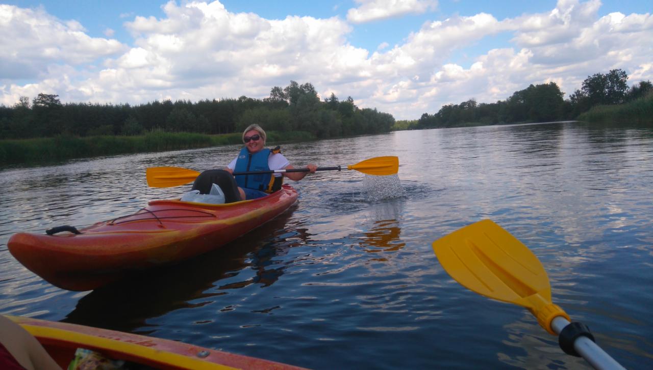 Uśmiechnięta kobieta w kamizelce asekuracyjnej płynie kajakiem po spokojnej rzece, wiosło w ruchu tworzy bryzę wody, w tle zielone drzewa i błękitne niebo z białymi chmurami.
