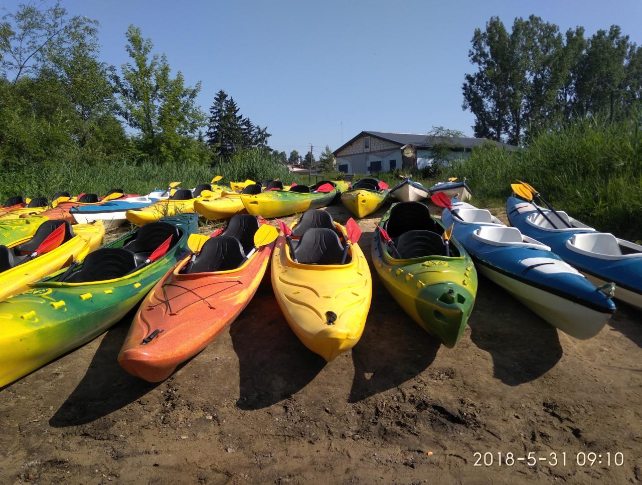 Różnokolorowe kajaki z wiosłami ułożone na ziemi, przygotowane do spływu; w tle budynek i zieleń.