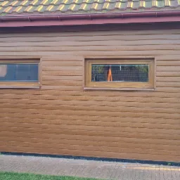 Elewacja budynku obłożona brązowym sidingiem z dwoma małymi oknami, widok na podwórko z ozdobnym grillem i lampami.