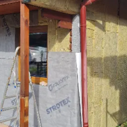Budynek w trakcie ocieplania elewacji: widoczna warstwa izolacji z wełny mineralnej, folia paroizolacyjna z nadrukiem 'STROTEX' i 'FOLIOREX', drewniana stolarka okienna i drzwiowa, oraz fragment...