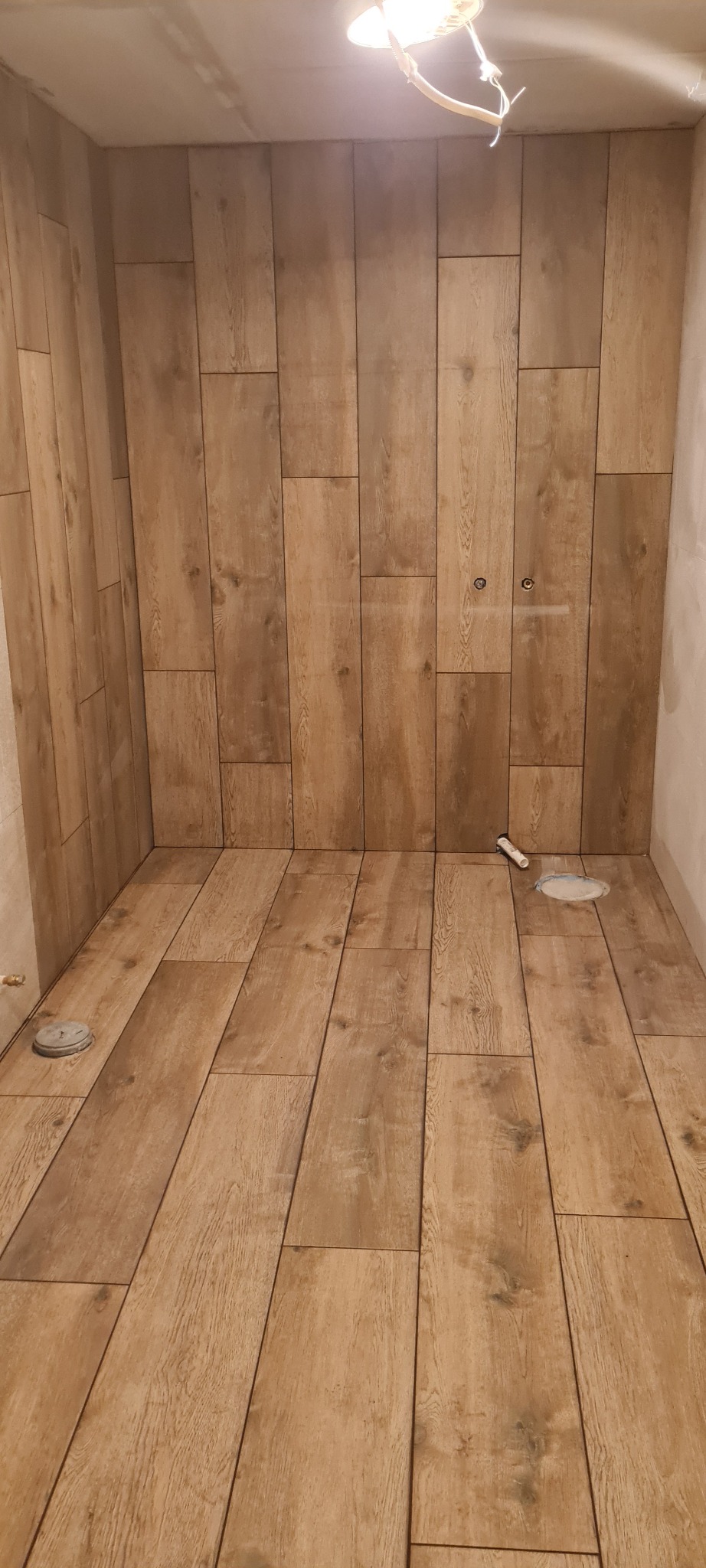 Wykończona łazienka z płytkami imitującymi drewno na podłodze i ścianach, widoczne otwory na armaturę i niedokończone podłączenie oświetlenia sufitowego.