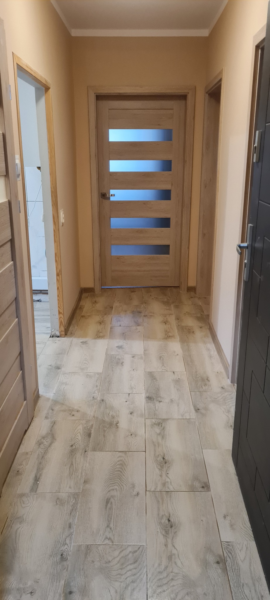 Wykończony korytarz z jasnymi płytkami imitującymi drewno, beżowe ściany i drewniane drzwi z przeszkleniami, widok na otwarte przejście do innego pomieszczenia.