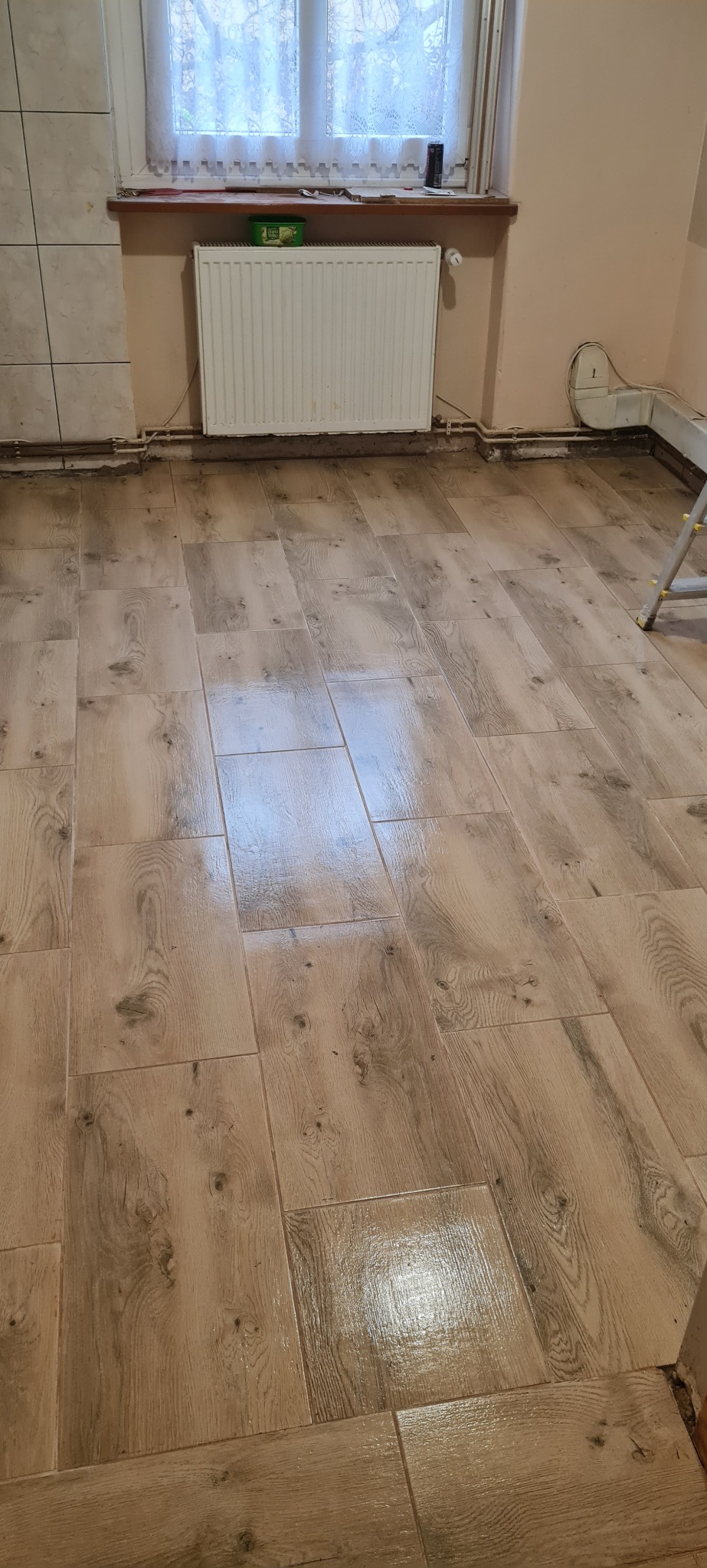 Ułożona podłoga z płytek imitujących drewno w jasnym odcieniu, widoczny grzejnik pod oknem oraz fragment drabiny malarskiej w rogu pomieszczenia.