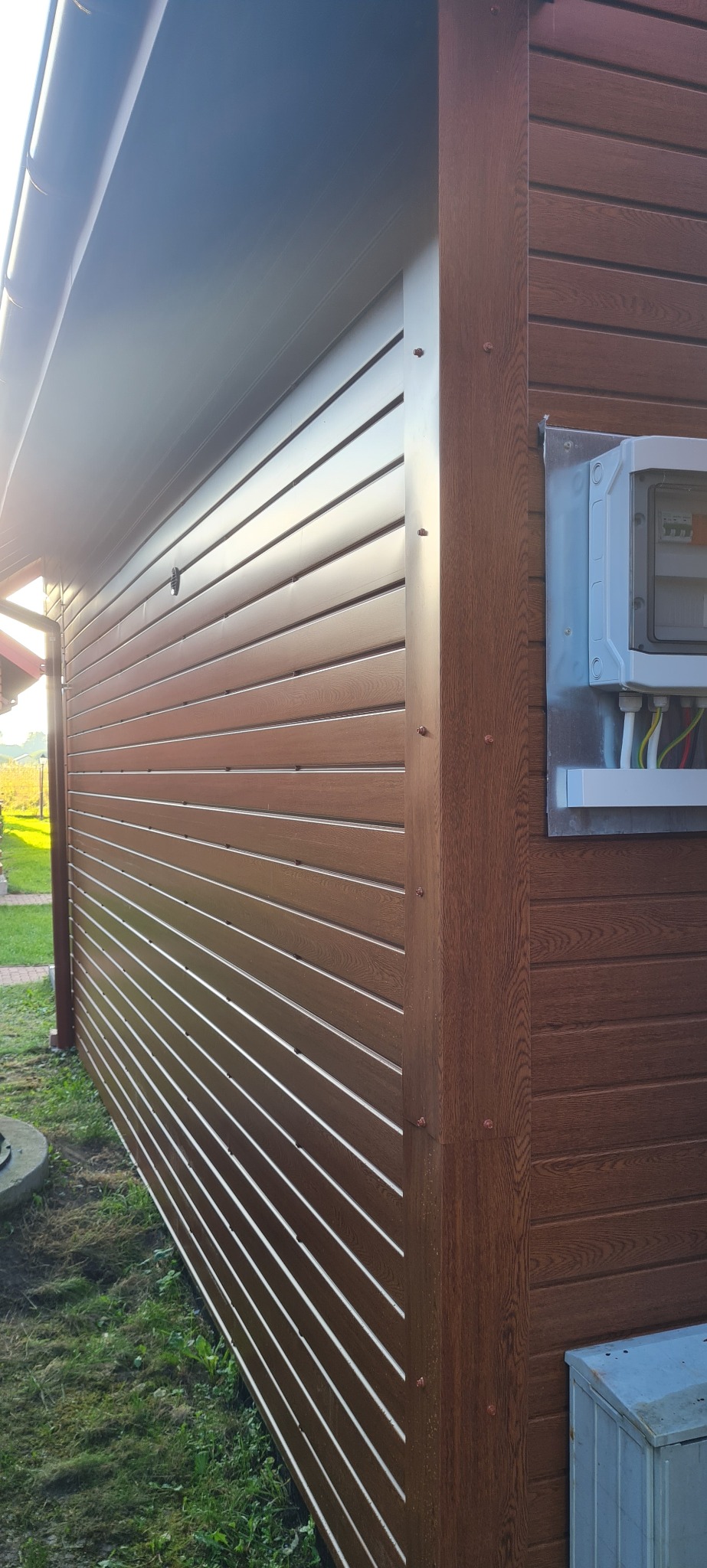 Brunatna elewacja budynku z panelami imitującymi drewno, widoczna skrzynka elektryczna i narożnik z wyraźną strukturą drewna.