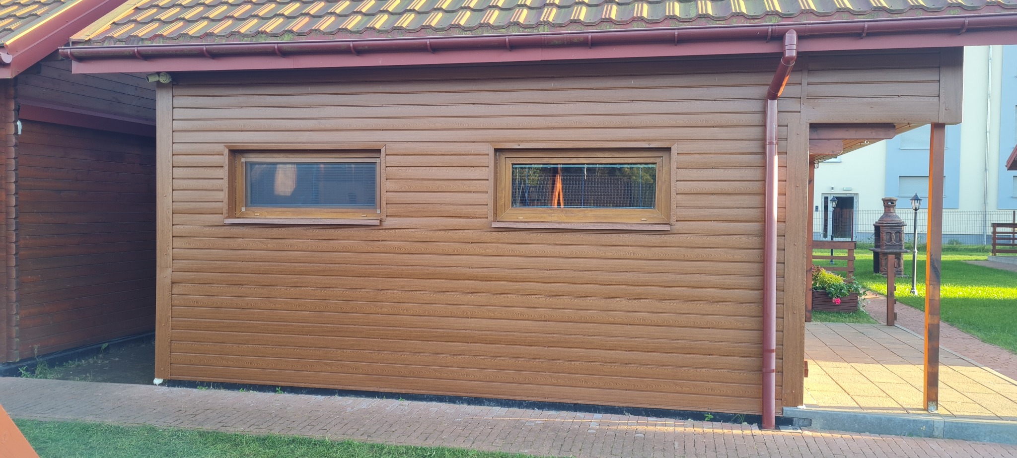 Elewacja budynku obłożona brązowym sidingiem z dwoma małymi oknami, widok na podwórko z ozdobnym grillem i lampami.