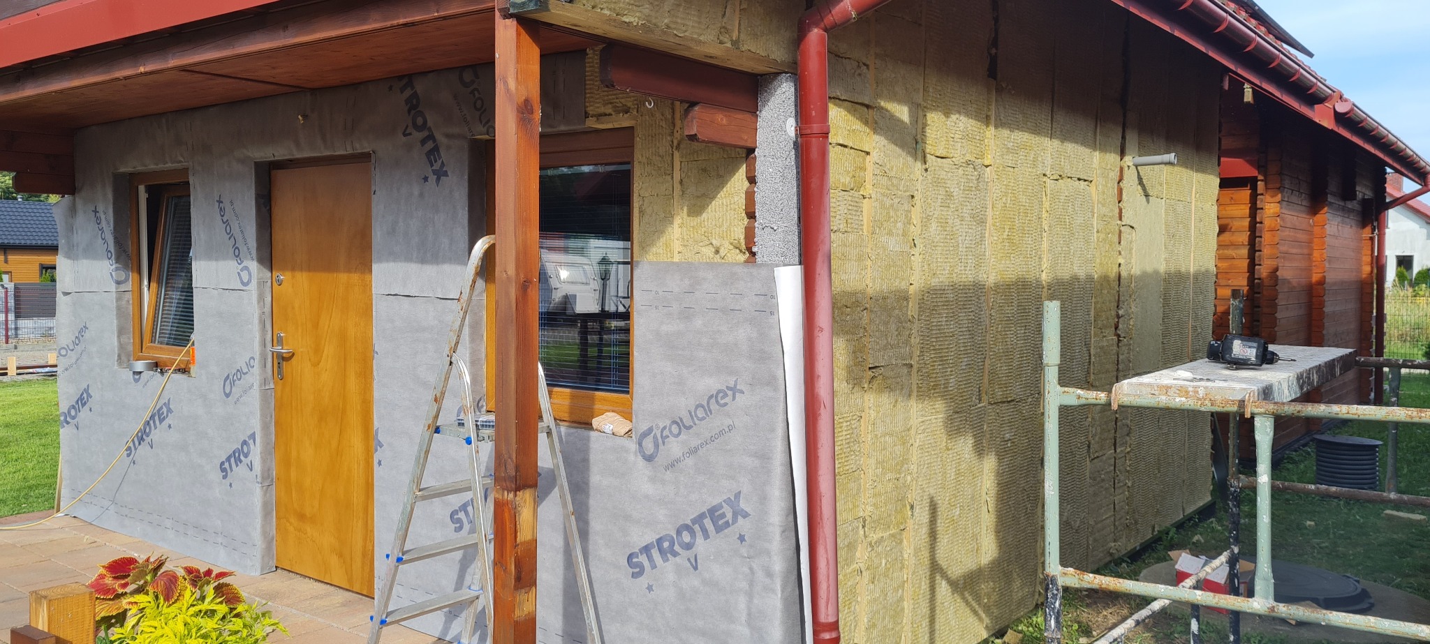 Budynek w trakcie ocieplania elewacji: widoczna warstwa izolacji z wełny mineralnej, folia paroizolacyjna z nadrukiem 'STROTEX' i 'FOLIOREX', drewniana stolarka okienna i drzwiowa, oraz fragment...