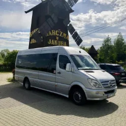Srebrny bus Mercedes-Benz Sprinter z przyciemnianymi szybami zaparkowany na brukowanym placu przed drewnianym wiatrakiem z napisem 'Karczma u Jakuba'.