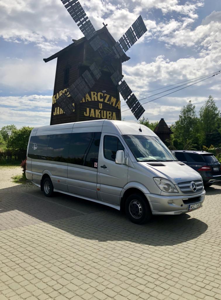 Srebrny bus Mercedes-Benz Sprinter z przyciemnianymi szybami zaparkowany na brukowanym placu przed drewnianym wiatrakiem z napisem 'Karczma u Jakuba'.