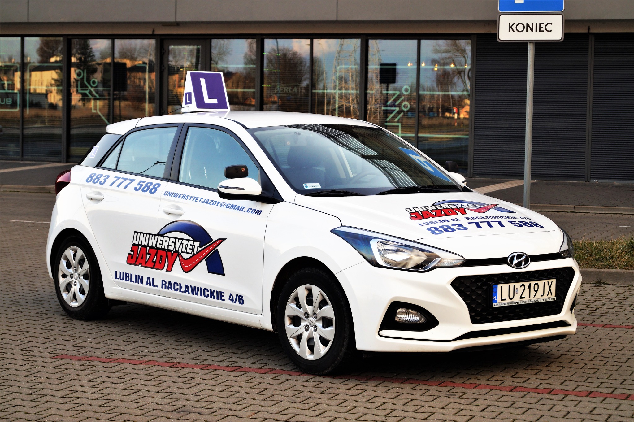 Biały samochód z oznaczeniem 'L' na dachu, zaparkowany na chodniku z kostki, z logo i numerem telefonu na karoserii, na tle nowoczesnego budynku biurowego.