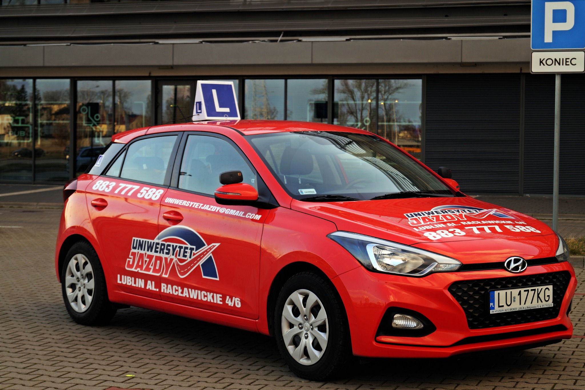 Czerwony samochód z oznaczeniem 'L' na dachu, zaparkowany na brukowanej nawierzchni, z widocznymi danymi kontaktowymi i logo na karoserii.