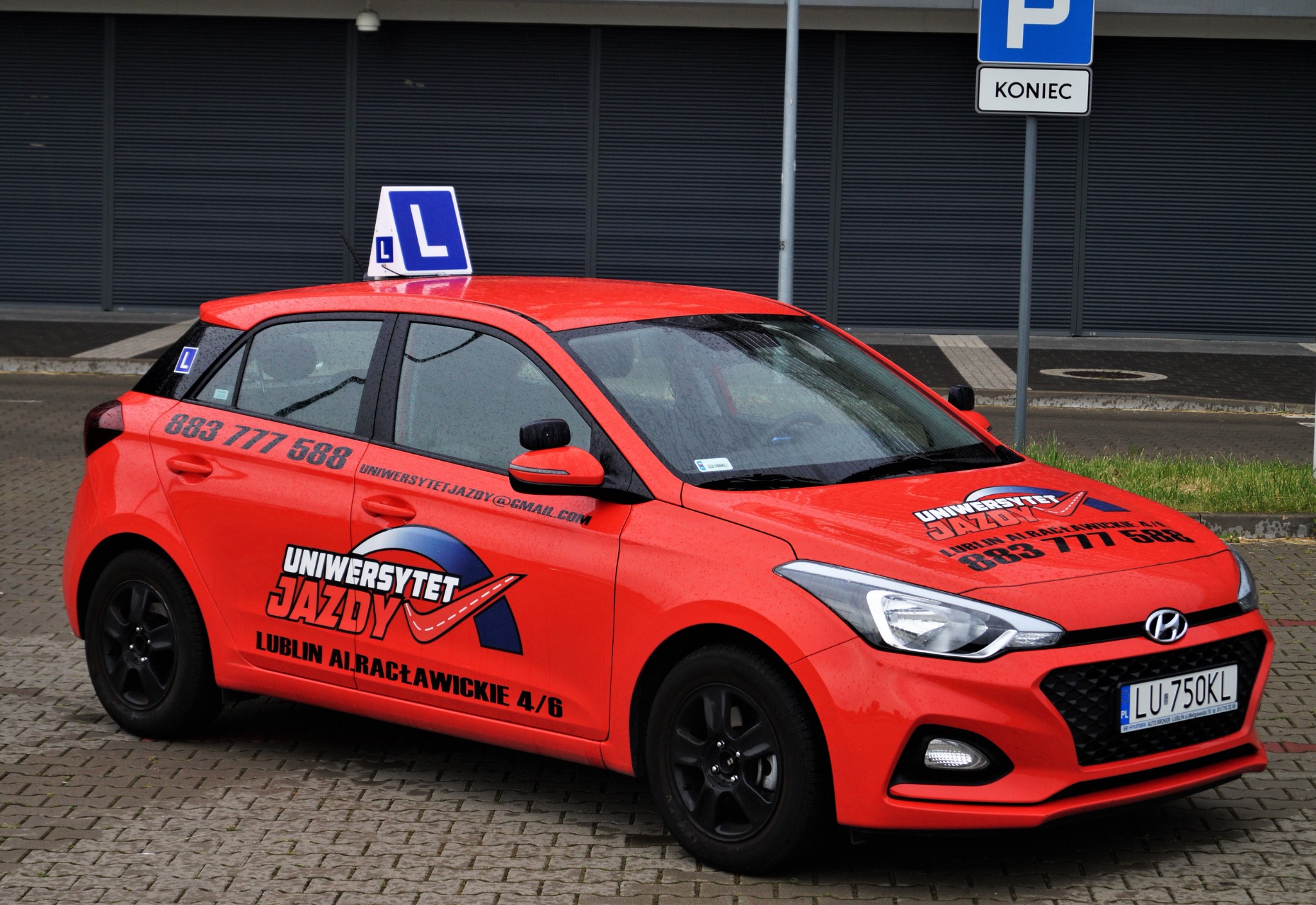 Czerwony samochód oznaczony literą 'L' na dachu, z logo i danymi kontaktowymi 'Uniwersytet Jazdy' na karoserii, zaparkowany na brukowanej nawierzchni przed szarą ścianą.