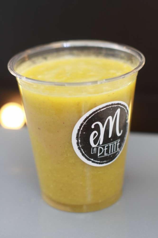 Żółty smoothie w przezroczystym kubku z naklejką 'em LA PETITE', na szarym blacie, z rozmytym światłem w tle.