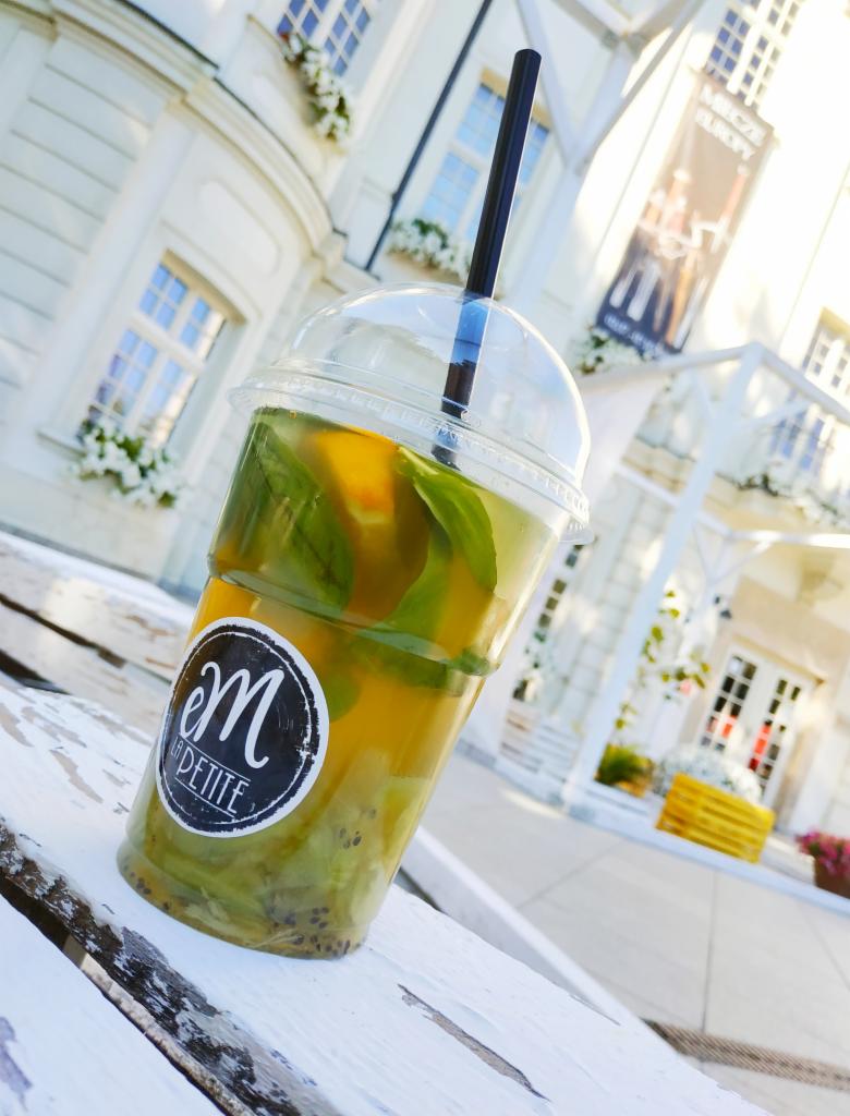 Orzeźwiający napój z kiwi, pomarańczą i miętą w przezroczystym kubku z czarną słomką, na tle białej, stylowej architektury z kwiatami w oknach.