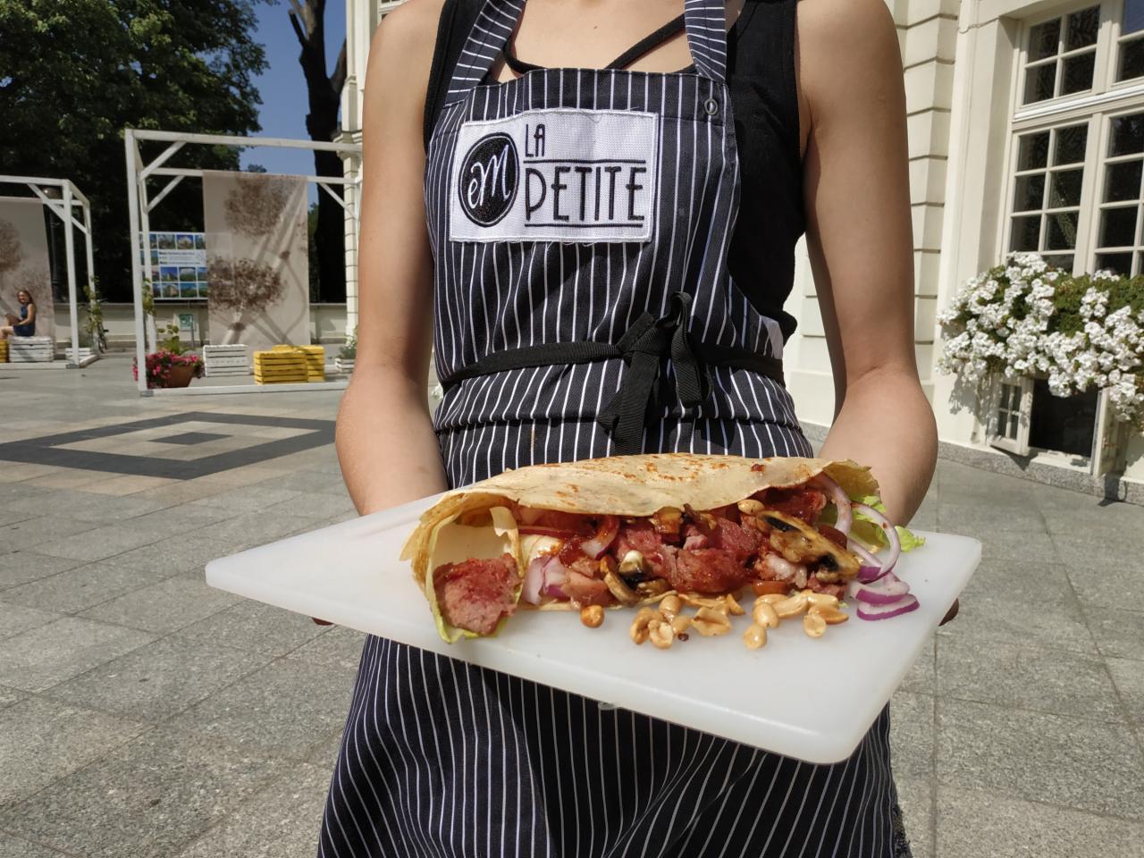 Osoba w pasiastej zapasce z logo 'La Petite' prezentuje naleśnik zawijany z mięsem, pieczarkami, czerwoną cebulą i orzechami na białej desce do krojenia na tle brukowanej nawierzchni i budynku...