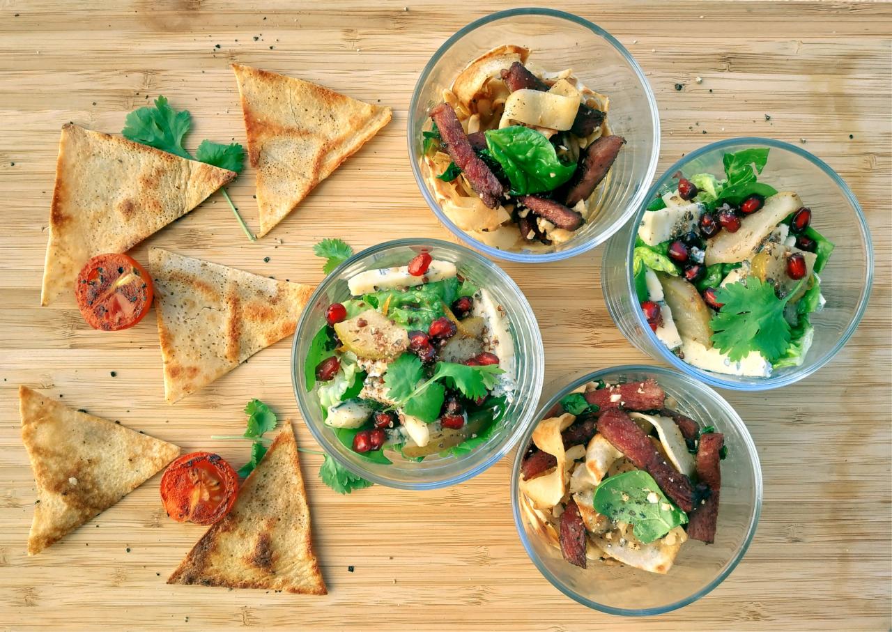 Kompozycja z czterech szklanych misek z sałatkami i trójkątnych pieczonych chlebków pita oraz pomidorów na drewnianej desce.