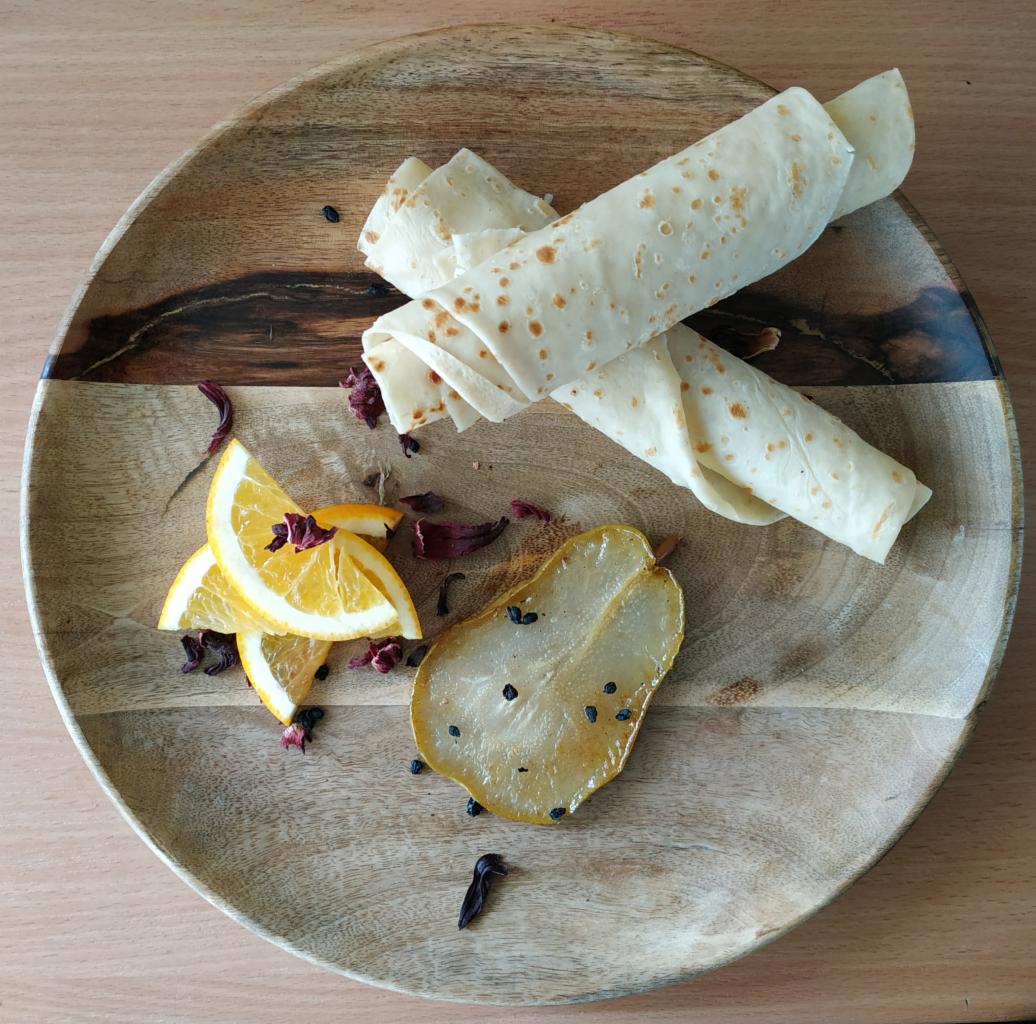 Dwa rulony naleśników na drewnianym talerzu z plasterkami pomarańczy, karmelizowaną gruszką i suszonymi kwiatami hibiskusa.