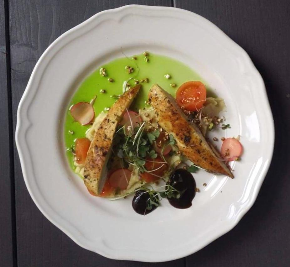 Elegancko podane danie na białym talerzu: dwa filety z kurczaka ułożone na puree, z dodatkiem rzodkiewek, pomidorów cherry, zielonego sosu i ciemnego balsamicznego glazuru, udekorowane mikrolistkami.