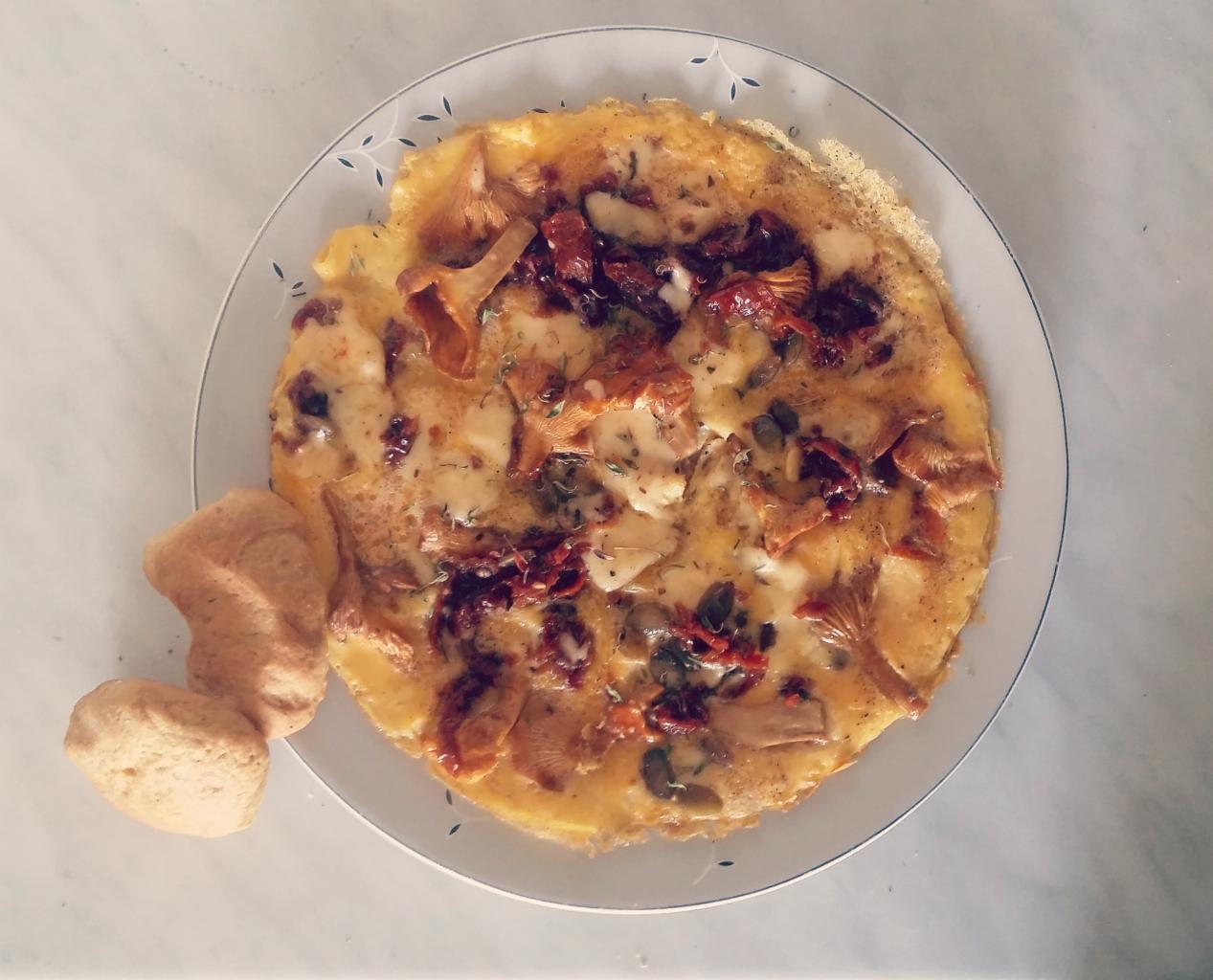 Omlet z kurkami, suszonymi pomidorami i ziołami na talerzu z niebieskim wzorem, obok dwa kawałki pieczywa.