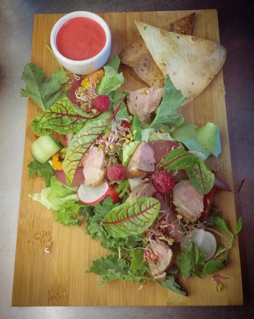 Sałatka z plastrami pieczonego mięsa, malinami, rzodkiewką i kiełkami, podana na drewnianej desce z sosem w białej miseczce i trójkątami pieczonego ciasta.