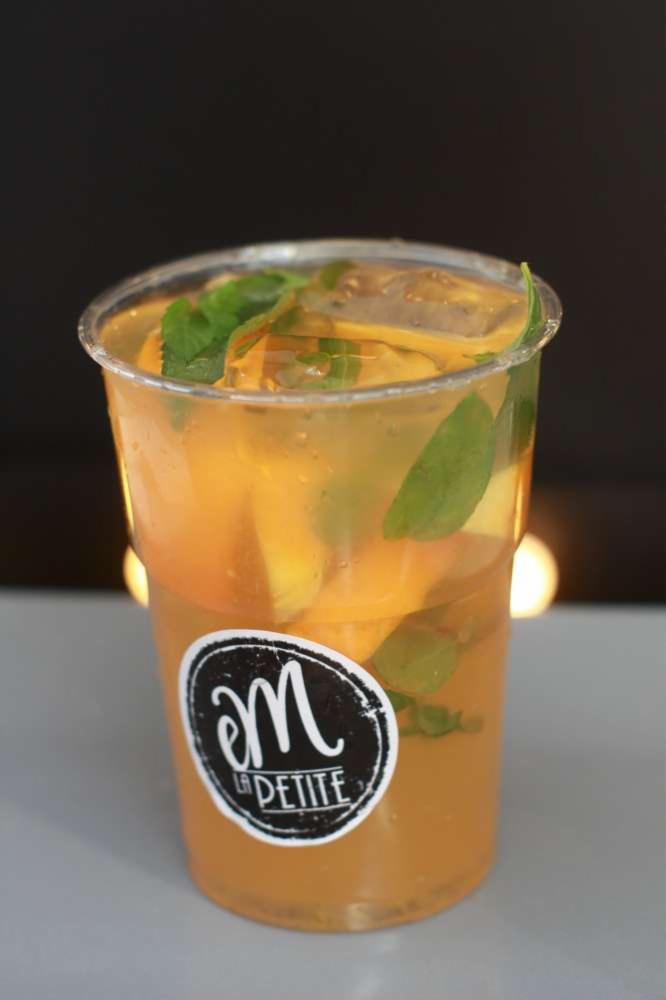 Napój pomarańczowy z miętą i lodem w przezroczystym plastikowym kubku z logo 'm La Petite' na etykiecie.
