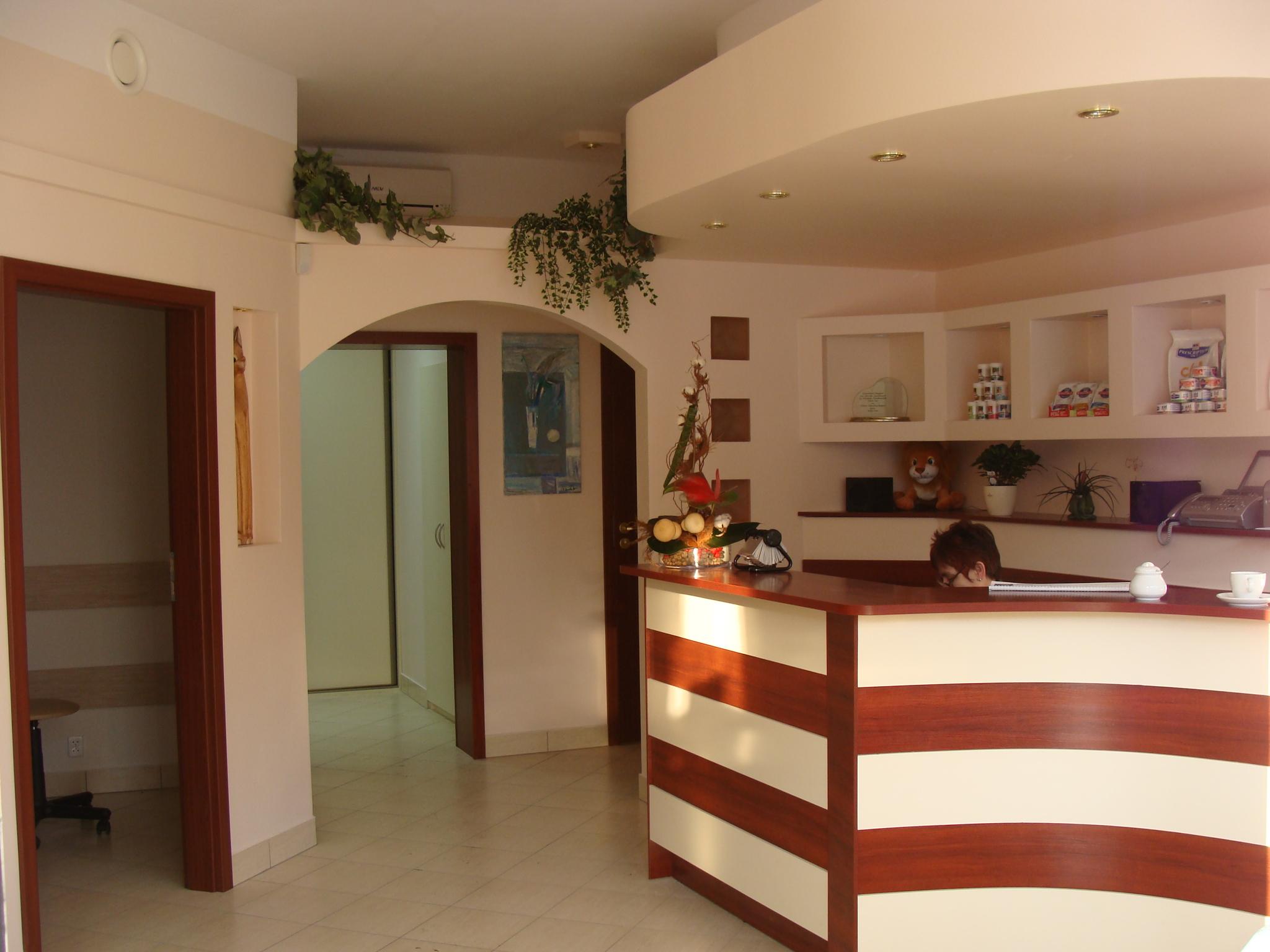 Recepcja biurowa z ladą w brązowo-białe pasy, dekoracjami roślinnymi na ścianach i suficie, oraz widocznym fragmentem osoby za ladą.