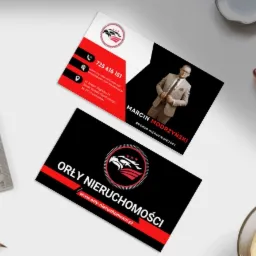 Dwie wizytówki firmy Orły Nieruchomości leżące na biurku obok kawy, ołówka i linijki. Górna wizytówka z numerem telefonu, adresem e-mail i zdjęciem Marcina Modrzyńskiego, brokera nieruchomości.