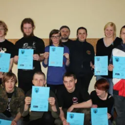 Grupa osób, część siedząca, część stojąca, prezentuje niebieskie certyfikaty z godłem na tle beżowej ściany.