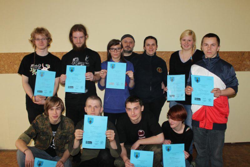 Grupa osób, część siedząca, część stojąca, prezentuje niebieskie certyfikaty z godłem na tle beżowej ściany.