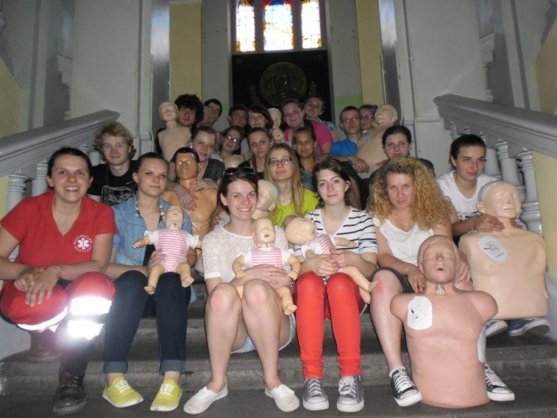 Grupa osób, siedząca na schodach, trzymająca manekiny do ćwiczeń resuscytacji, w tym manekiny niemowląt.